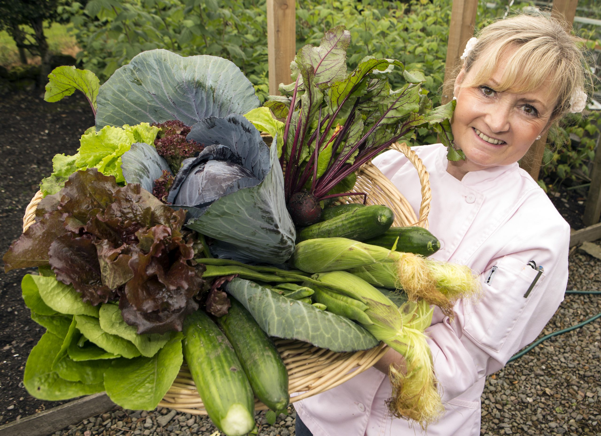 Woman holding a box of fresh vegetables and salad with a market garden behind.